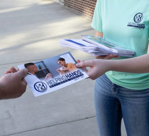 Person handing out "Helping Hands" flyers on a sidewalk, wearing a light blue Helping Hands t-shirt.