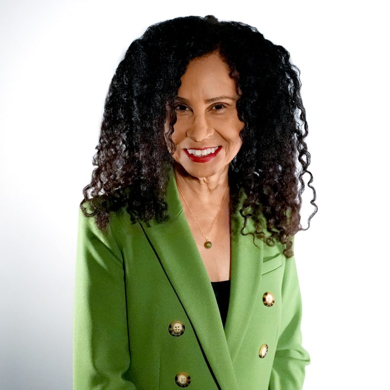 Smiling woman with curly hair in a green blazer, standing against a plain white background.