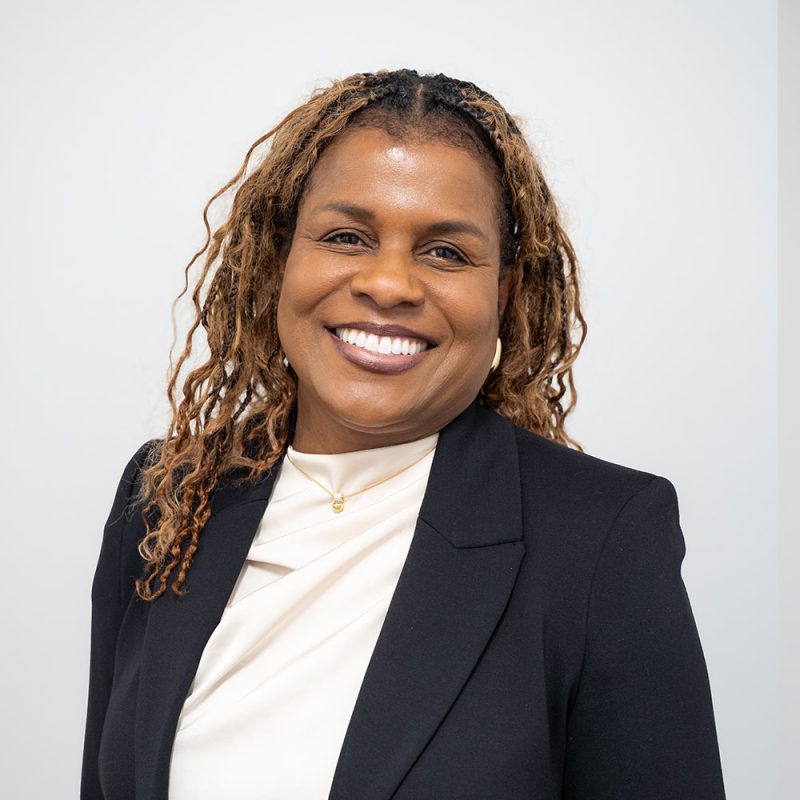 Smiling woman with long curly hair in a black blazer and white top against a plain light background.