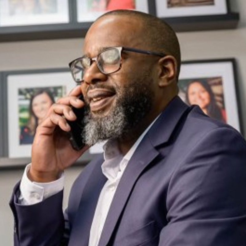 Man in a suit with glasses and a beard talking on the phone, smiling, with framed photos in the background.