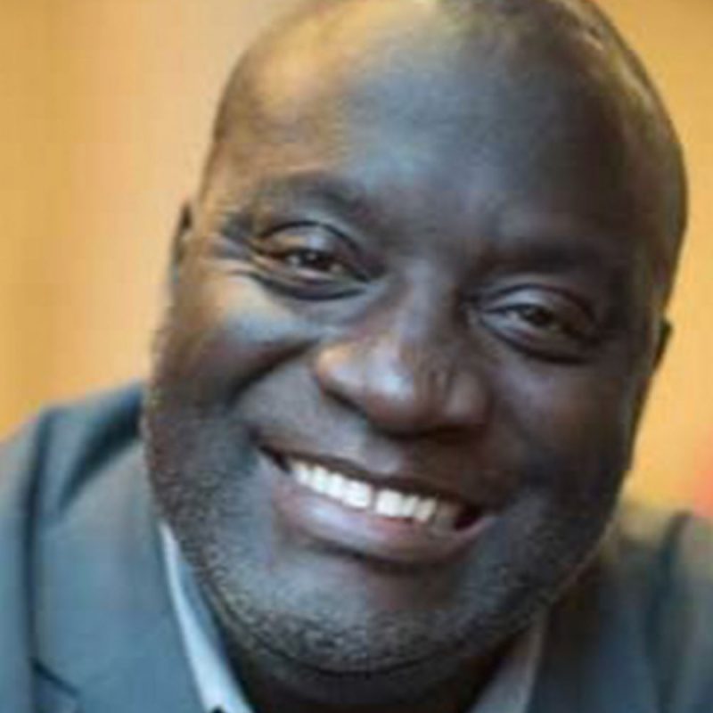 A man in a suit smiling warmly at the camera, with a blurred background.
