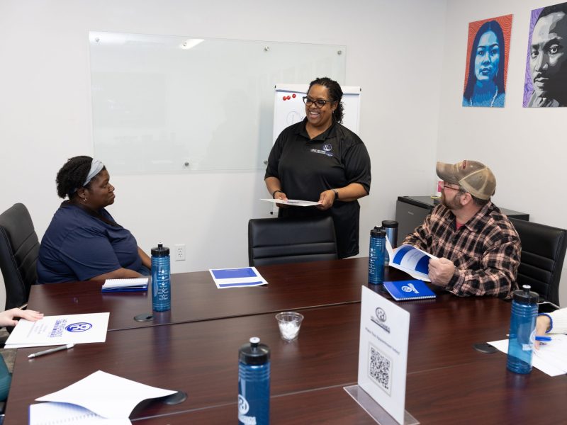 Four people sit around a conference table while a woman stands and speaks, holding papers in a meeting room.
