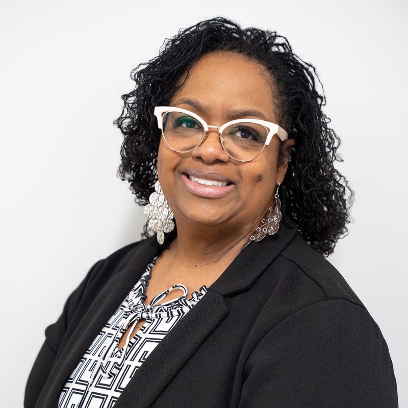 Smiling woman with curly hair, glasses, and earrings, wearing a black blazer and patterned blouse.